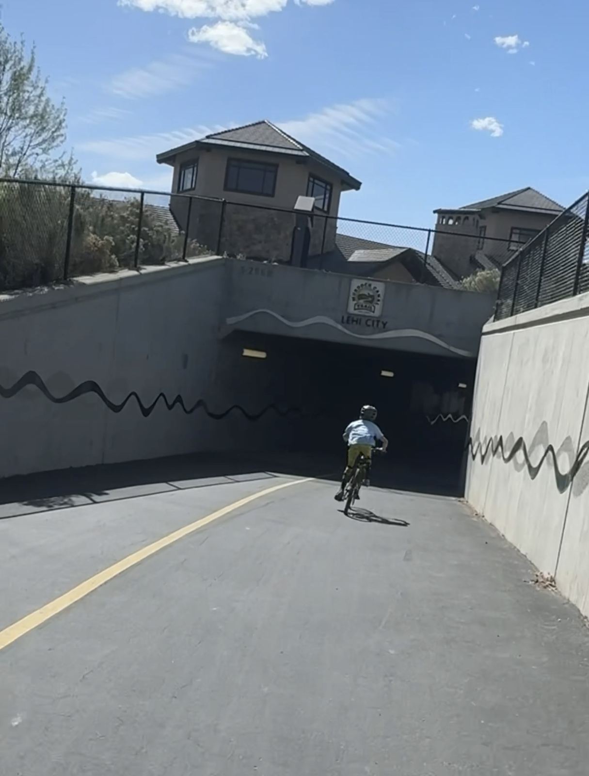 Child on a bike, seen from behind, riding down the paved Murdock Canal Trail into a concrete underpass marked "Lehi City" under a clear blue sky.