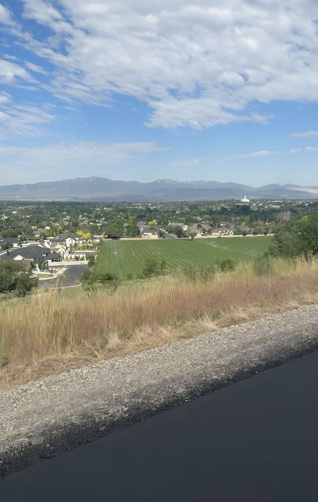 Freshly paved trail edge with dry grass in the foreground, a green field and homes below, and a mountain range with a small white spired building on the horizon under scattered clouds.