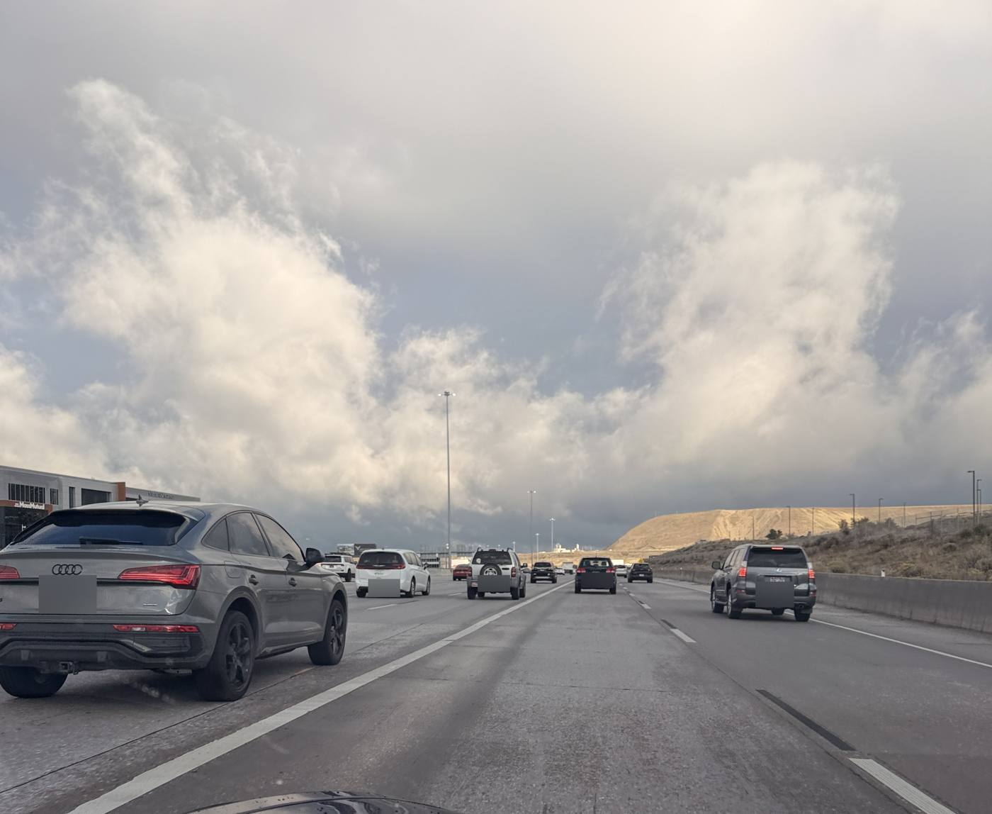 Rush hour traffic on a multi lane Utah County freeway under heavy gray storm clouds, with a tan hillside on the right