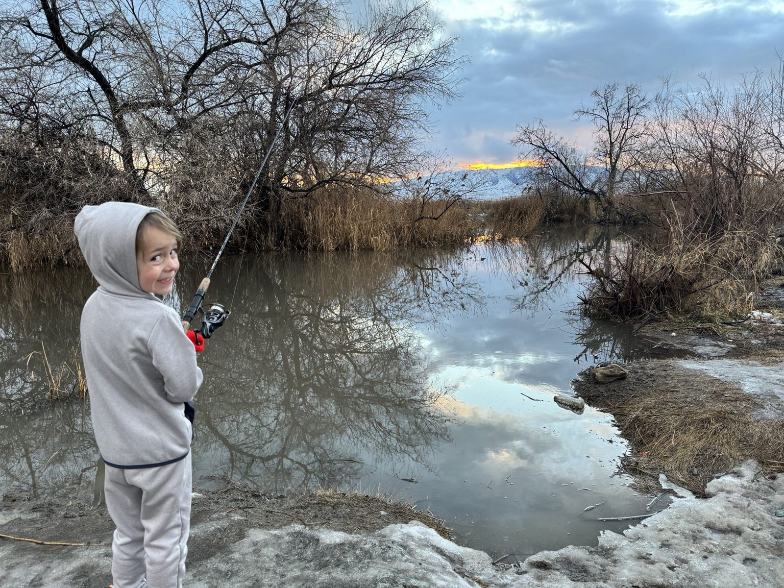 Child in a gray hooded sweatsuit holds a fishing rod and looks back over one shoulder, standing on a snowy, muddy bank beside a still creek that reflects bare trees, dry reeds, and a cloudy sky glowing orange near snow covered mountains.