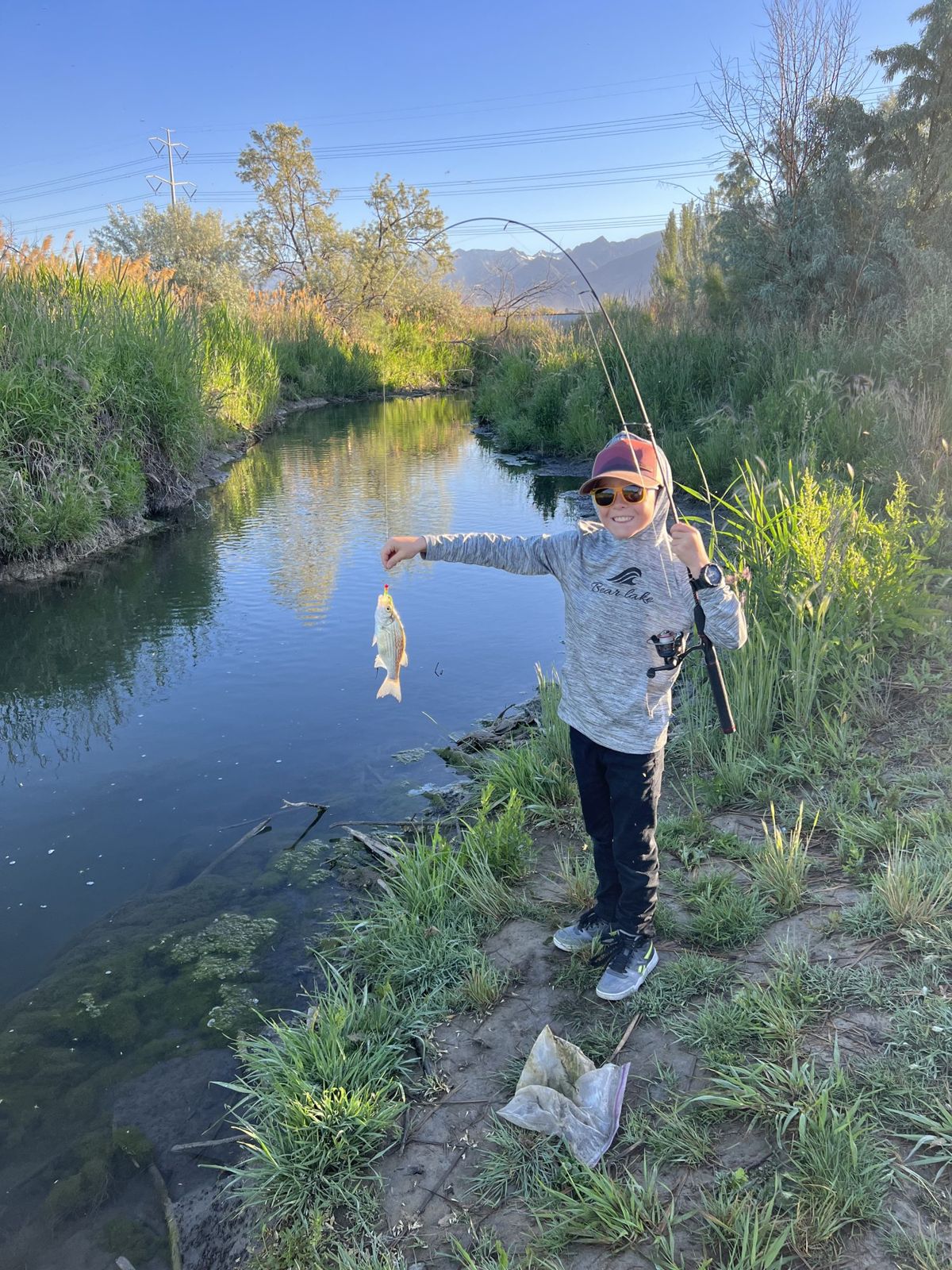 Smiling child in a red cap, sunglasses, and a gray hoodie stands on a grassy bank holding a fishing rod and a small silvery fish on the line, beside a calm creek lined with green reeds, with mountains in the distance under a clear blue sky.